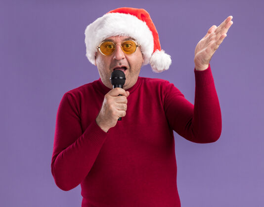 年龄困惑的中年男子戴着圣诞老人帽 戴着黄色眼镜 举着胳膊站在紫色背景上对着麦克风说话举起圣诞节中间