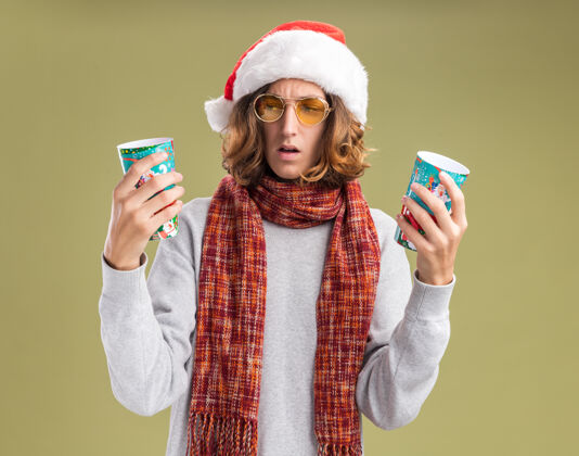 站着戴着圣诞老人帽 戴着黄色眼镜 脖子上围着暖和的围巾的年轻人手里拿着五颜六色的纸杯 站在绿色的背景上 满脸疑惑圣诞老人杯子围巾