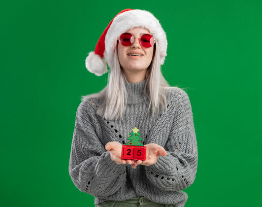 举行年轻的金发女郎穿着冬季毛衣 戴着圣诞帽 戴着红眼镜 手里拿着玩具立方体 圣诞节日期快乐而积极地看着相机 站在绿色的背景下微笑着眼镜立方体日期