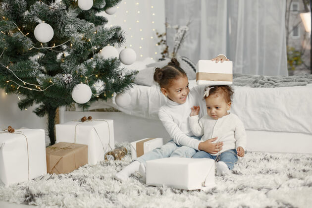 室内穿着白色毛衣的孩子女儿们在圣诞树旁缝线两个姐妹在家节日盒子年轻