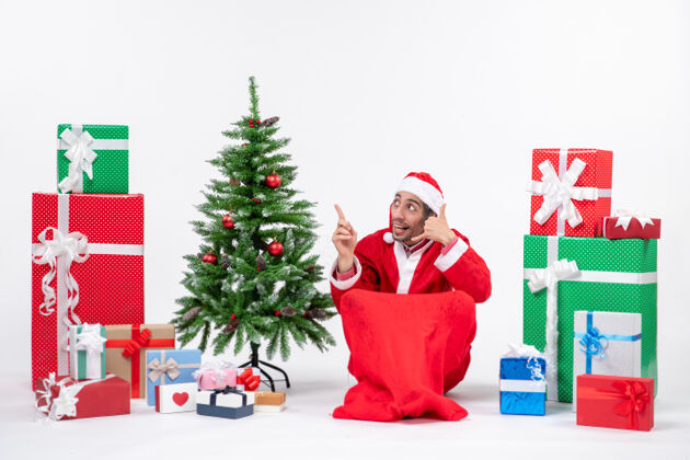坐着年轻人打扮成圣诞老人 拿着礼物和装饰好的圣诞树坐在地上做着叫我的手势 指着上面庆祝地圣诞老人