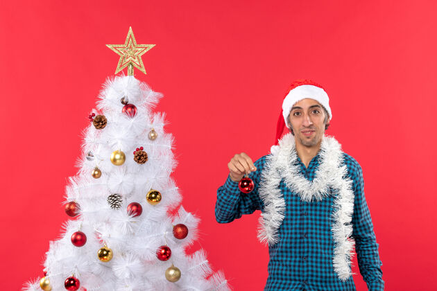 庆祝滑稽的年轻人戴着圣诞老人的帽子 穿着蓝色的衬衫 手里拿着装饰品圣诞老人年轻人配件