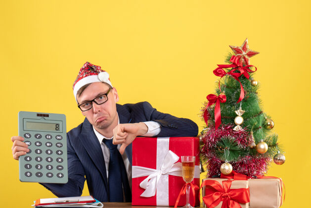 圣诞节前视图沮丧的商人与圣诞树附近的圣诞帽坐在桌上 并在黄色的礼物公司新郎圣诞树