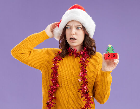 斯拉夫震惊的年轻斯拉夫女孩戴着圣诞帽 脖子上戴着花环 手拿圣诞树饰物 看着紫色背景上孤立的一面 还有复制空间装饰品举行年轻