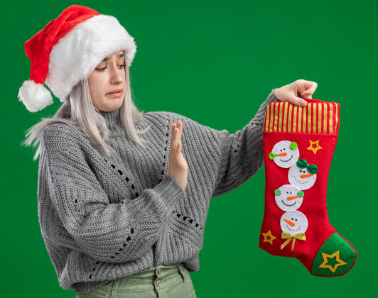 长袜年轻的金发女郎穿着冬衣 戴着圣诞帽 手里拿着圣诞长袜 站在绿色的背景上 做着一个防御的手势 看上去既困惑又担心使手金发