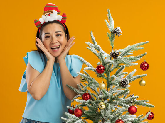 边缘身穿蓝色上衣的年轻女子头戴有趣的圣诞环 看着相机 高兴而兴奋地站在橙色背景下的圣诞树旁树有趣头