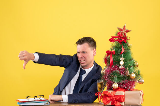 行政人员愤怒的年轻人坐在圣诞树旁的桌子旁做着拇指朝下的手势 黄色的墙上挂着礼物年轻人生意人
