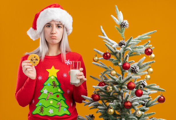 帽子穿着圣诞毛衣 戴着圣诞帽 手里拿着一杯牛奶和饼干 表情悲伤的金发女郎毛衣看着橙色
