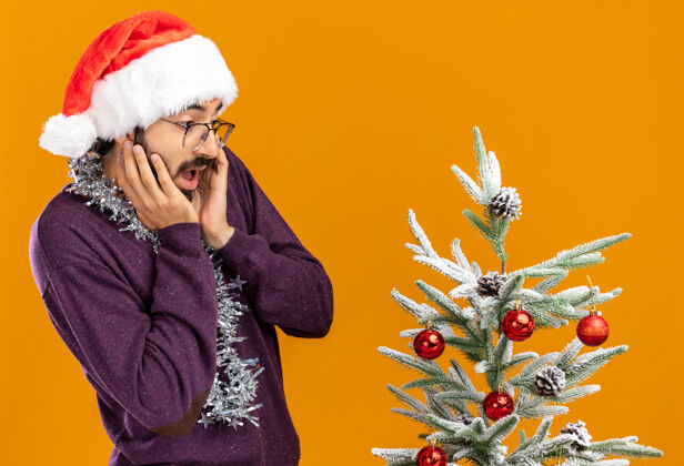 惊讶惊讶的年轻帅哥站在圣诞树旁 戴着圣诞帽 脖子上戴着花环 手放在脸颊上 橘黄色的背景隔离开来帽子圣诞树