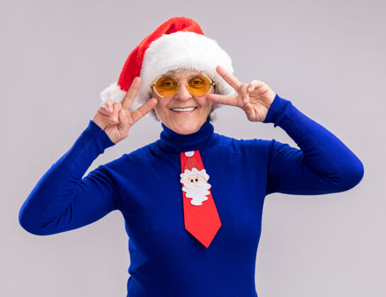 手势戴着太阳眼镜 戴着圣诞帽 系着圣诞领带 面带微笑的老妇人 在白色背景上用复制空间孤立地打着胜利的手势胜利微笑眼镜