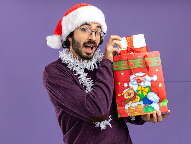 礼物微笑着的年轻帅哥戴着圣诞帽 脖子上戴着花环 手里拿着蓝色背景的礼品袋脖子帽子圣诞节