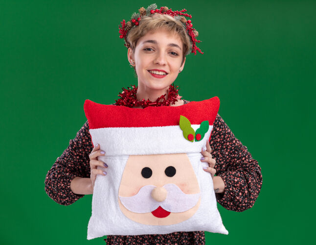 头微笑着的年轻漂亮女孩戴着圣诞花环 脖子上戴着金箔花环 抱着圣诞老人的枕头 看着隔离在绿色背景上的相机枕头圣诞老人漂亮