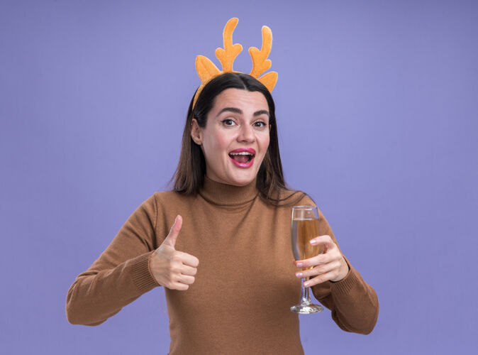 微笑微笑着的年轻漂亮女孩 穿着棕色毛衣 戴着圣诞发箍 手里拿着一杯香槟 在蓝色的背景上孤立地竖起大拇指篮球拇指女孩