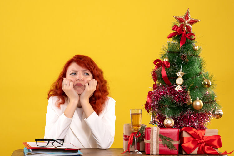 女性女工人坐在桌子后面 手里拿着圣诞礼物和黄色的圣诞树礼物黄色桌子