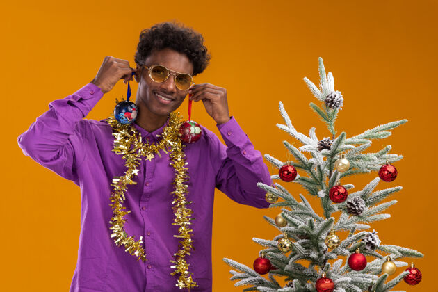 圣诞节微笑着的年轻的非裔美国人戴着眼镜 脖子上戴着金属丝花环 站在装饰过的圣诞树旁 把圣诞饰品挂在耳朵上 孤立地挂在橙色的墙上悬挂花环年轻