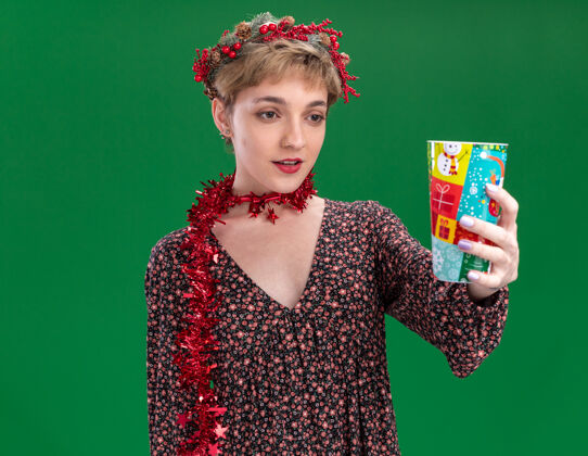 朝向给人印象深刻的年轻漂亮女孩戴着圣诞花环 脖子上戴着金属丝花环 伸出塑料圣诞杯 看着隔离在绿色墙上的它花环塑料周围