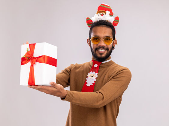 脸一个穿着棕色毛衣 头戴圣诞老人耳环 打着有趣的红色领带的非裔美国人拿着礼物 站在白色背景下 面带微笑地看着相机领带轮辋微笑