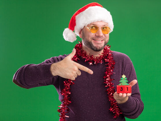 金属片微笑着的中年男子戴着圣诞帽 脖子上戴着金箔花环 戴着眼镜 拿着圣诞树玩具看 日期看着隔离在绿色背景上的相机帽子脖子中年
