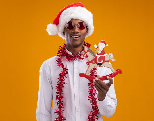 印象深刻年轻的美国黑人男子戴着圣诞帽 戴着眼镜 脖子上戴着金箔花环 抱着摇摇木马上的圣诞老人 看着橙色背景上的相机脖子金属片非裔美国人