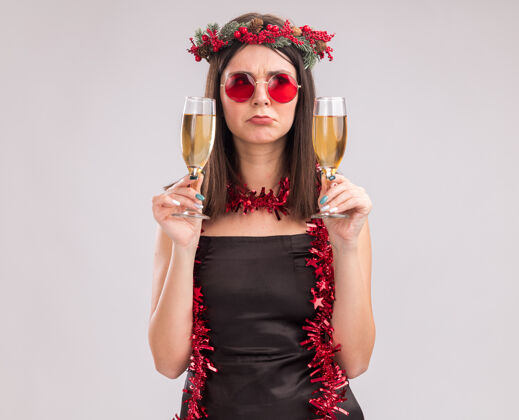 周围困惑的年轻漂亮的白人女孩戴着圣诞花环 脖子上戴着金箔花环 戴着一副眼镜 手里拿着两杯香槟花环复制圣诞
