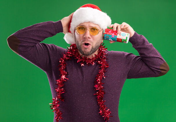 圣诞快乐好奇的中年男子戴着圣诞帽 脖子上戴着金属丝花环 戴着眼镜 手里拿着塑料圣诞杯 耳边听着秘密秘密保持快乐