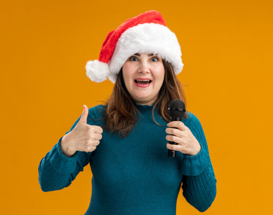 帽子兴奋的成年白人妇女 戴着圣诞帽 竖起大拇指 拿着麦克风隔离在橙色背景上 留有复制空间拇指圣诞老人坚持