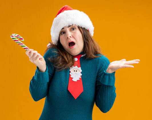 帽子困惑的成年白人妇女 戴着圣诞帽 打着圣诞领带 拿着糖果手杖 被隔离在橙色的背景上 还有复制空间橙色手杖成人