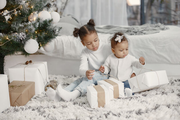 盒子穿着白色毛衣的孩子女儿们在圣诞树旁缝线两个姐妹在家女儿室内毛衣