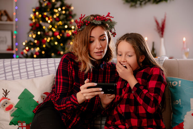 妈妈令人印象深刻的母亲和女儿坐在沙发上看电话 享受圣诞节的时间在家里印象深刻电话一些东西
