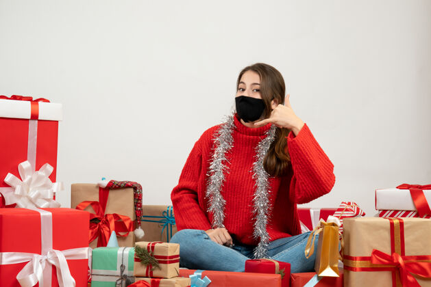 年轻女孩穿着红色毛衣 戴着黑色面具的年轻女孩在做“打电话给我”的手势 围坐在白色的礼物旁边坐着圣诞节电话