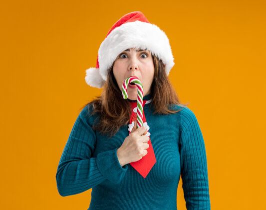 橘子惊讶的成年白人妇女戴着圣诞帽 打着圣诞领带 手里拿着糖果棒 橙色背景上有复制空间大人领带抱着