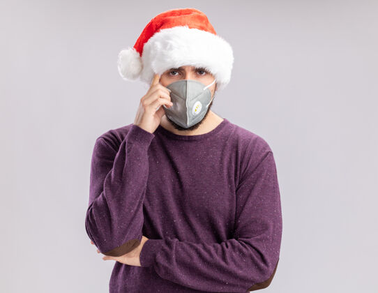 圣诞老人一个穿着紫色毛衣 戴着圣诞帽 戴着护面面具的年轻人站在白色背景下 严肃地看着镜头严肃站着保护