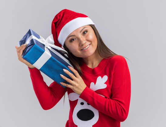 脸微笑着歪着头的年轻亚洲女孩戴着圣诞帽和毛衣拿着礼品盒 脸上孤立着白色的背景倾斜圣诞节围着