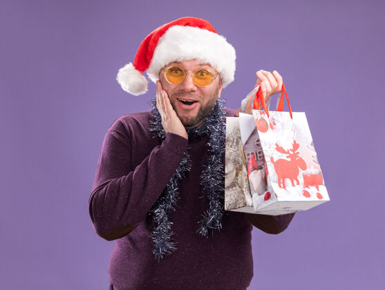 脸兴奋的中年男子戴着圣诞帽 脖子上戴着金属丝花环 戴着眼镜 手里拿着圣诞礼品袋 看着相机 手放在脸上 在紫色的背景下与世隔绝兴奋帽子手