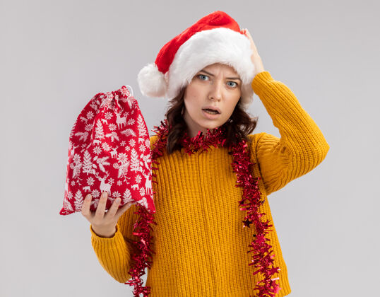 圣诞老人焦虑的年轻斯拉夫女孩戴着圣诞帽 脖子上戴着花环 手放在头上 手拿着圣诞礼品袋 背景是白色的 有复印空间斯拉夫周围年轻