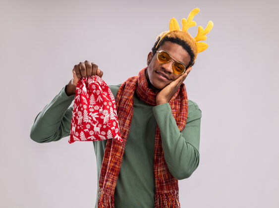 圣诞老人一个非洲裔美国人 戴着有趣的边 戴着鹿角 脖子上围着围巾 手里拿着红色的圣诞老人包 手里拿着礼物 站在白色的背景下 他开心而积极地微笑着角微笑鹿