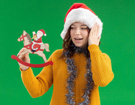 摇摆兴奋的年轻斯拉夫女孩戴着圣诞帽 脖子上戴着花环 抱着圣诞老人 看着摆在摇马上的圣诞老人持有绿色兴奋