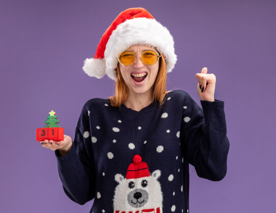 手势兴奋的年轻漂亮女孩穿着圣诞毛衣戴着圣诞帽戴着眼镜拿着圣诞玩具在紫色的墙上显示“是”的手势毛衣年轻穿着