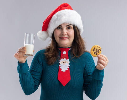 大人戴着圣诞帽 打着圣诞领带 拿着一杯牛奶和饼干 在白色背景上隔离开来 并留有复印空间的成年白种女人很高兴抱着高兴杯子