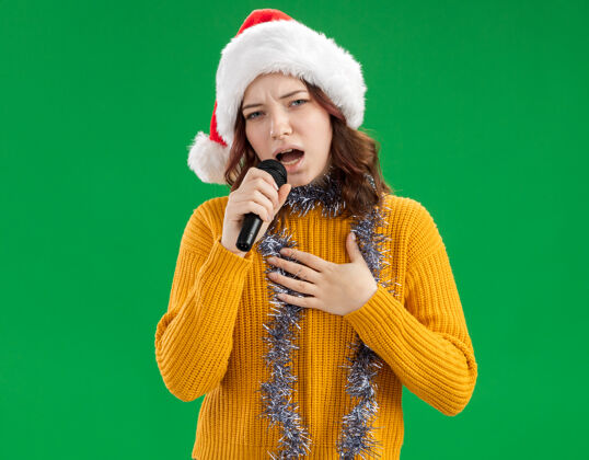 唱歌自信的年轻斯拉夫女孩戴着圣诞帽 脖子上戴着花环 手里拿着麦克风 假装在绿色背景上唱着与世隔绝的歌 还有复制空间周围麦克风帽子