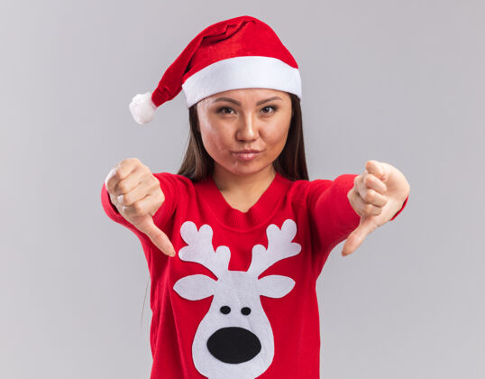 拇指高兴的年轻亚洲女孩戴着圣诞帽和毛衣显示大拇指向下孤立的白色背景高兴帽子年轻