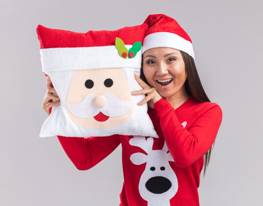 女孩Joyfyl年轻的亚洲女孩戴着圣诞帽 穿着毛衣 抱着白色背景的圣诞枕头枕头年轻毛衣