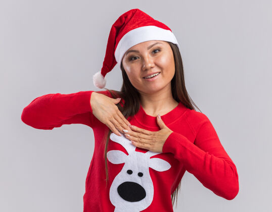 年轻高兴的年轻亚洲女孩戴着圣诞帽和毛衣把双手放在自己孤立的白色背景高兴推杆女孩