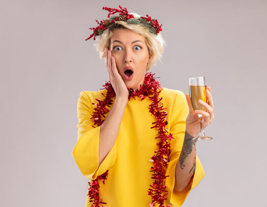 花环关心的金发女郎戴着圣诞花环 脖子上戴着金箔花环 手里拿着一杯香槟 手放在脸上 隔离在白色的墙上 留着复印空间玻璃穿着金箔
