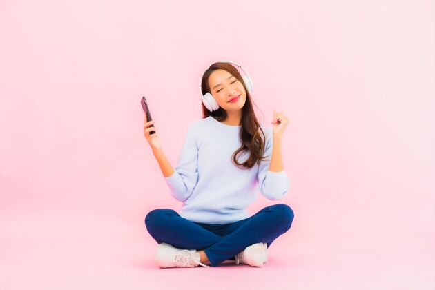 和谐肖像美丽的亚洲年轻女子用智能手机和耳机在粉红色的墙上听音乐明亮舞者黑发