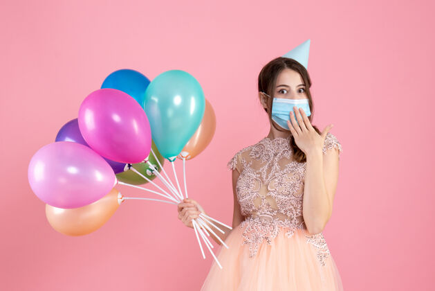 生日好奇的女孩戴着派对帽 手放在嘴上 手里拿着粉色的彩色气球举行帽子派对