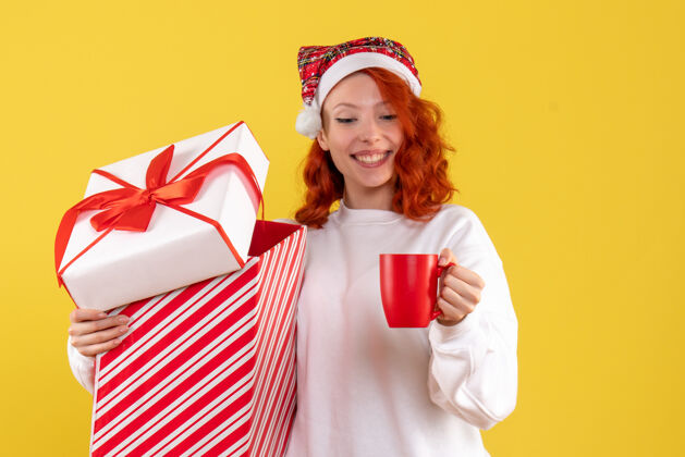 茶黄色墙上年轻女子手持圣诞礼物和一杯茶的正面图微笑快乐人