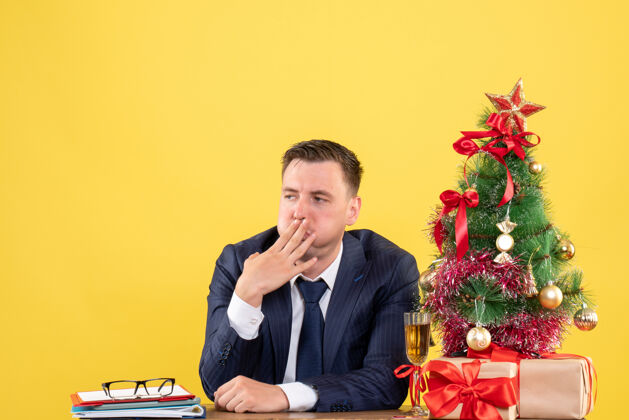 人不满意的男人坐在圣诞树旁的桌子前 呈现黄色圣诞节不满意行政人员