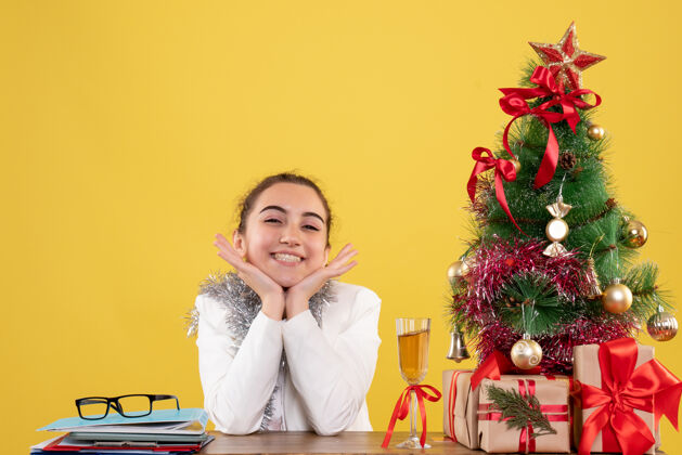 情绪正面图：坐在桌子后面的女医生兴奋地站在黄色背景上 手里拿着圣诞树和礼品盒成人人圣诞节