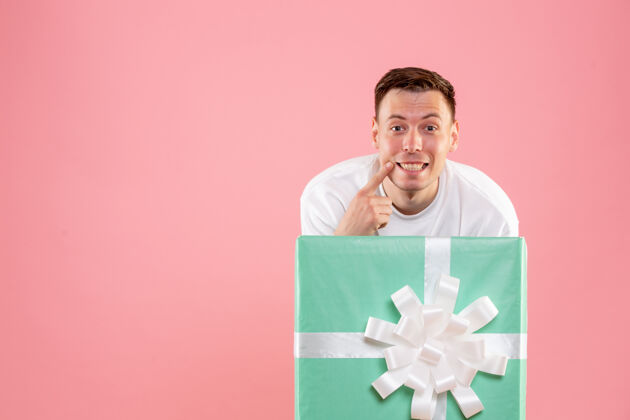 礼品盒隐藏在礼物里的年轻人在粉红色墙上微笑的正面视图庆祝圣诞快乐聚会